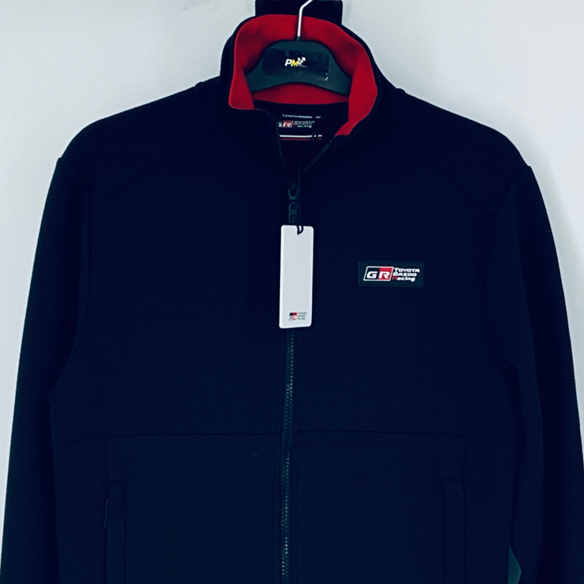 Dark navy full-zip sweatshirt with red-lined hood, chest logo patch, hanging white tag