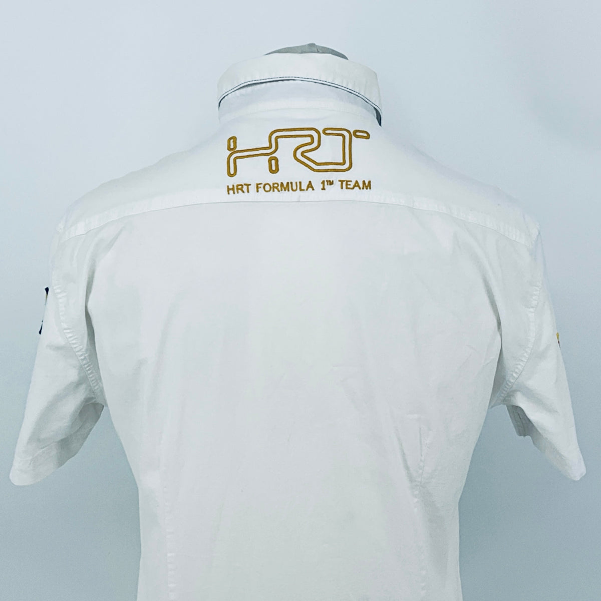 White pit crew shirt back with embroidered “HRT FORMULA 1 TEAM” logo across the shoulders.