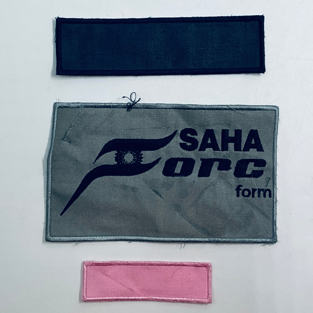 Three rectangular embroidered patches: dark blue bar, gray SAHA ORCA logo, and pink strip.