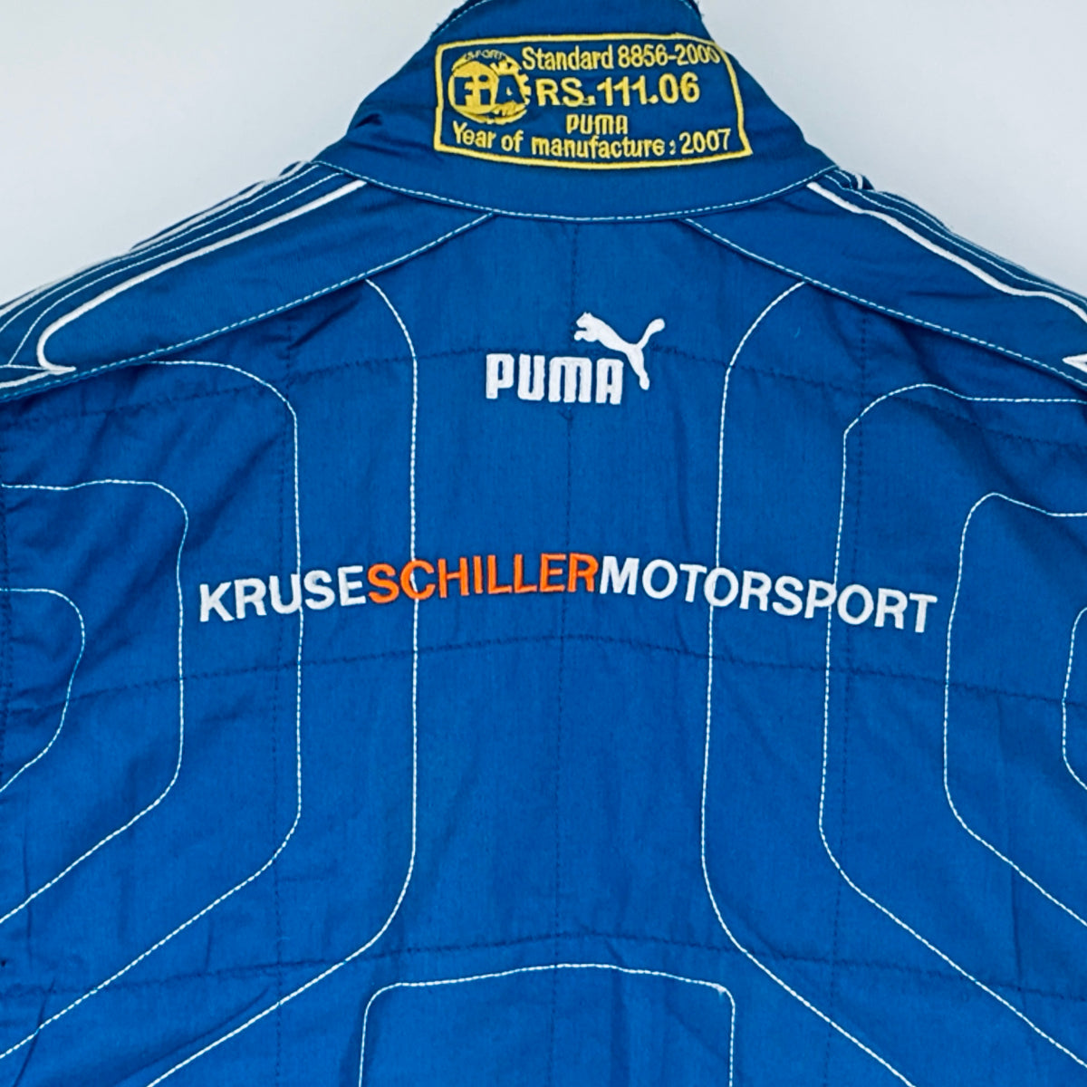 Blue driver suit collar label with FIA text, Puma logo, and KRUSE SCHILLERMOTORSPORT